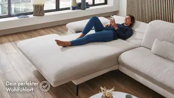 Frau liegt entspannt auf ausziehbarer heller Stoffcouch in modernem Wohnzimmer mit großen Fenstern und Holzfußboden – zeigt den Komfort und die Funktionalität der Couch als gemütlichen Rückzugsort.